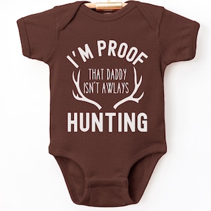 May include: A brown baby onesie with white text that reads "I'm proof that daddy isn't always hunting" with a white antler graphic.