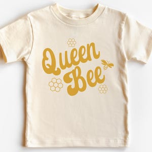 May include: A cream colored t-shirt with the text "Queen Bee" in a yellow retro font. The text is surrounded by small yellow honeycomb designs and a yellow bee.