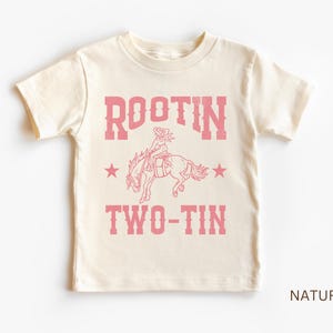 May include: A white t-shirt with a pink graphic of a cowgirl riding a horse. The graphic says "Rootin' Two-Tin" with stars on either side.