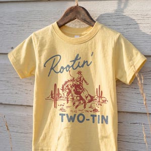 May include: A pale yellow short-sleeve t-shirt featuring a dark red graphic of a cowboy riding a horse in a desert landscape with cacti. The text Rootin' is printed in dark blue above the graphic, and the text TWO-TIN is printed below in blue.
