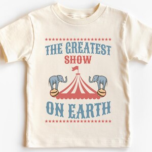 May include: A white t-shirt with a vintage circus theme design. The text reads "The Greatest Show On Earth" with a red and white striped tent and two elephants balancing balls.