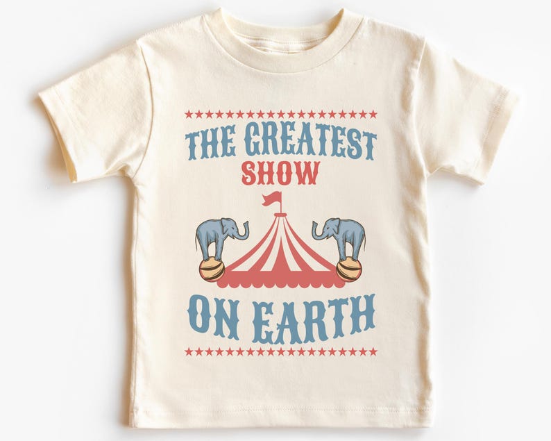 May include: A white t-shirt with a vintage circus theme design. The text reads "The Greatest Show On Earth" with a red and white striped tent and two elephants balancing balls.
