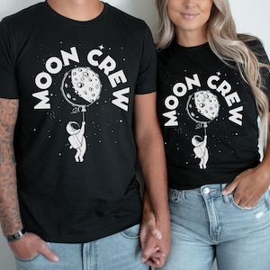 May include: Two black t-shirts with the text "Moon Crew" in white. The shirts feature a white astronaut holding a moon with a black crescent shape.