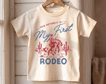 Puede incluir: Una camiseta de manga corta color crema con un estampado gráfico en rojo y azul. El texto dice "***THIS ACTUALLY IS*** My First RODEO" sobre una ilustración de un vaquero montando un caballo en un paisaje desértico con cactus.