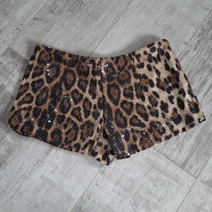 May include: A pair of leopard print shorts with a sequined finish. The shorts feature a brown and black spotted pattern, with small silver sequins scattered across the fabric. The shorts are made of a soft material.