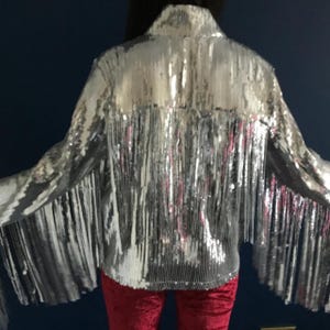 May include: Silver sequin jacket with fringe detail. The jacket has long sleeves and a collared neckline. The fringe hangs from the back and sleeves. The model is wearing red velvet pants. The jacket is a statement piece.