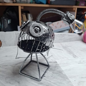 May include: A handcrafted, silver-colored metal anglerfish lamp. The lamp features a round body made of overlapping metal scales, a wireframe mouth, and a flexible neck supporting a lightbulb. It stands on a geometric base.