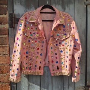 Può includere: Una giacca di jeans rosa decorata con strass colorati e borchie dorate. La giacca ha una chiusura a zip e maniche lunghe.