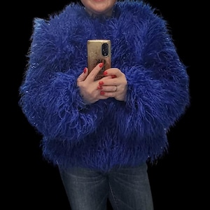 May include: A striking blue, fluffy jacket with a textured finish. The jacket features long sleeves and a round neckline. The person in the image is holding a gold-coloured phone. The person is wearing blue jeans.