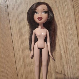 Könnte beinhalten: Eine nackte Plastikpuppe mit langen braunen Haaren und rotem Lidschatten. Die Puppe trägt eine rosa-rote Perlenkette.