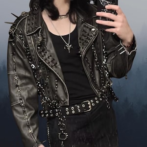 May include: A dark grey leather jacket, adorned with silver spikes, studs, and chains. The biker-style jacket features a notched collar, an asymmetrical zip, and a belt. The jacket has a distressed appearance.