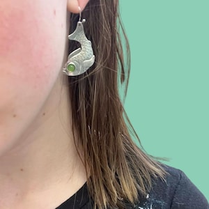 May include: Silver fish-shaped earrings with a green gemstone eye. The earrings feature scale and fin detailing, suspended from a silver hook. A unique and eye-catching accessory, perfect for adding a touch of whimsy.
