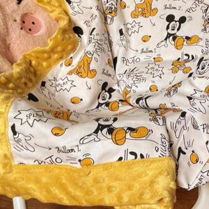 Pode incluir: Um cobertor de bebê branco com uma estampa de desenho animado do Mickey Mouse e Pluto e detalhes em amarelo. O design inclui as palavras "Balloon 7" e "Pluto". O cobertor tem uma borda amarela texturizada.