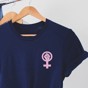 Puede incluir: Una camiseta azul marino con un s&iacute;mbolo feminista rosa con un pu&ntilde;o en alto.