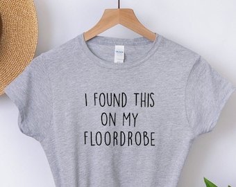 Op de afbeelding: Een lichtgrijs t-shirt met de zwarte tekst "I FOUND THIS ON MY FLOORDROBE". Het shirt hangt aan een houten hanger. Een strohoed is links te zien. Rechts staat een plant in een decoratieve pot.