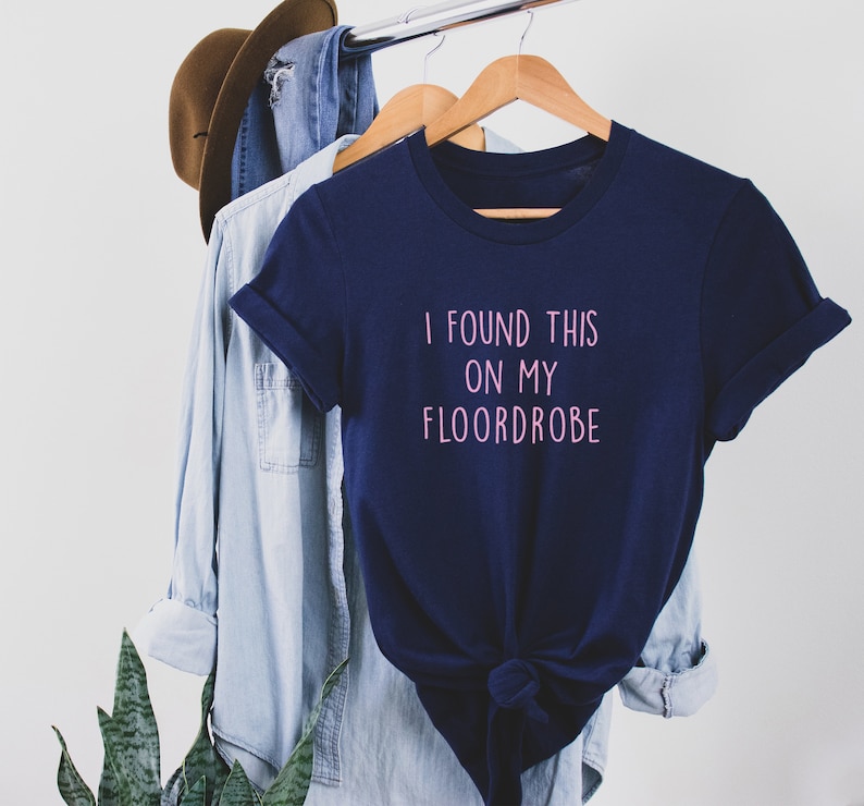 Puede incluir: Una camiseta azul marino con el texto "I found this on my floordrobe" en letras rosas.