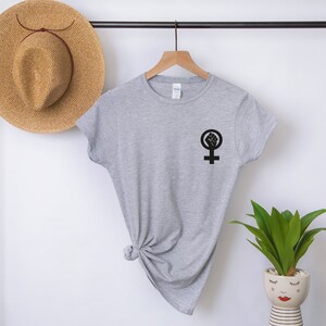 Puede incluir: Una camiseta gris con un s&iacute;mbolo feminista negro con un pu&ntilde;o levantado dentro del c&iacute;rculo.