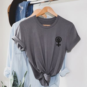 Puede incluir: Una camiseta gris con un s&iacute;mbolo feminista negro que presenta un pu&ntilde;o dentro de un c&iacute;rculo. El s&iacute;mbolo est&aacute; en el lado izquierdo de la camiseta.