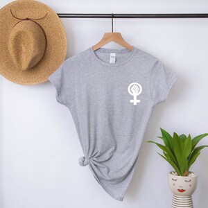 Puede incluir: Camiseta gris con un s&iacute;mbolo feminista blanco con un pu&ntilde;o dentro. El s&iacute;mbolo est&aacute; en el pecho izquierdo de la camiseta.