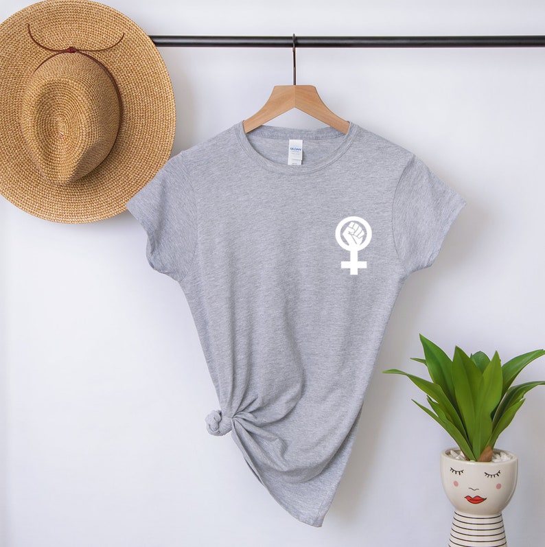 Puede incluir: Camiseta gris con un s&iacute;mbolo feminista blanco con un pu&ntilde;o dentro. El s&iacute;mbolo est&aacute; en el pecho izquierdo de la camiseta.