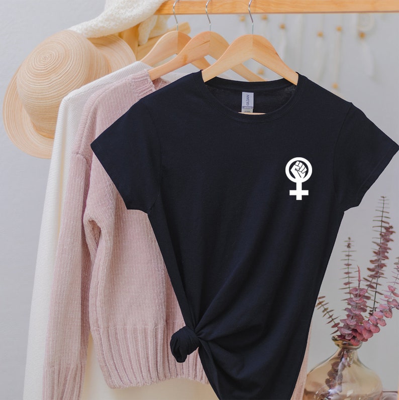 Puede incluir: Camiseta negra con un s&iacute;mbolo feminista blanco que presenta un pu&ntilde;o dentro de un c&iacute;rculo encima de un s&iacute;mbolo femenino.