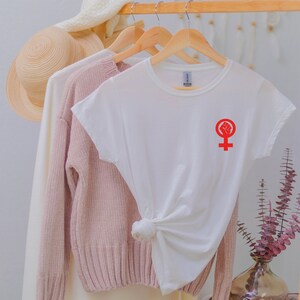 Puede incluir: Camiseta blanca con un s&iacute;mbolo feminista rojo con un pu&ntilde;o en alto. El s&iacute;mbolo est&aacute; en el lado izquierdo de la camiseta.