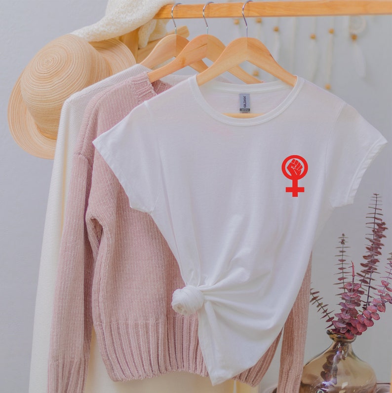 Puede incluir: Camiseta blanca con un s&iacute;mbolo feminista rojo con un pu&ntilde;o en alto. El s&iacute;mbolo est&aacute; en el lado izquierdo de la camiseta.