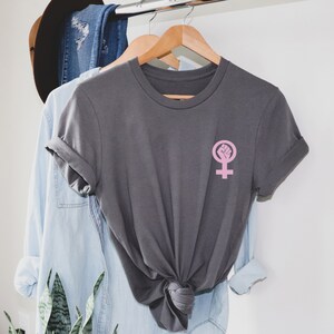 Puede incluir: Una camiseta gris antracita con un s&iacute;mbolo feminista rosa con un pu&ntilde;o en alto. La camiseta est&aacute; doblada y colgada en una percha con otras prendas.