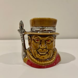 May include: A ceramic mug shaped like a jovial man wearing a top hat. The mug is primarily brown with a red collar and silver handle. The man's face has a smiling expression and red eyes.