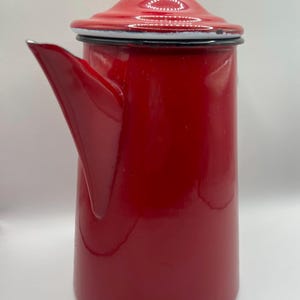 May include: A tall, red enamelware coffee pot with a lid and spout. The pot is a vibrant, glossy red with a black rim and handle. The lid has a small, round knob. A classic kitchenware item.
