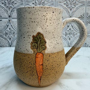 May include: A ceramic mug with a speckled off-white upper section and a speckled tan lower section. A hand-painted orange carrot with green leafy tops is centered on the front of the mug. The mug features a curved handle and a wide, rounded base.