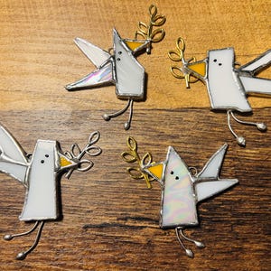 May include: Four stained glass dove ornaments. Each dove is white with iridescent wings, a yellow beak, and a gold-colored olive branch. The doves have silver wire legs and are set against a wooden background.