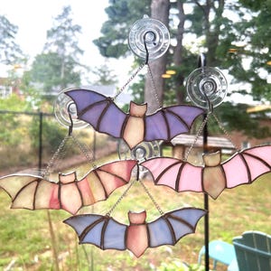 May include: Four stained glass bats hanging in a window. The bats are in shades of purple, pink, and yellow. They are suspended by chains and suction cups.