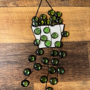 May include: A stained glass hanging featuring a white pot filled with green glass beads. The beads are strung on wire and hang down from the pot, creating a cascading effect.