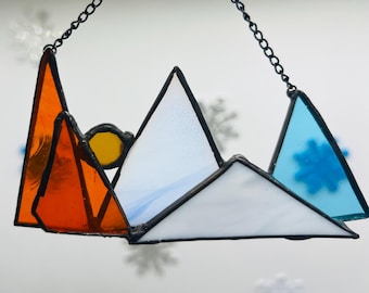 May include: A stained glass suncatcher featuring a mountain range with orange, white, and blue glass pieces. The suncatcher is suspended from a black chain.