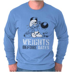 May include: Light blue long-sleeve shirt featuring a cartoon Smurf lifting weights. The Smurf is depicted in white and blue, with the text "WEIGHTS BEFORE DATES" in a distressed white and black font.