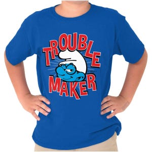 May include: A royal blue t-shirt featuring a cartoon Smurf character with the words "TROUBLE MAKER" in red block letters. The Smurf is blue with a white hat and a mischievous expression. The shirt is short-sleeved.