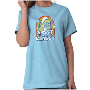 May include: Light blue t-shirt featuring a cartoon Smurfette character with a rainbow and the text "Sassy and Classy". The design includes hearts and a mirror. The shirt is a short-sleeved crew neck.
