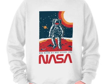 May include: White sweatshirt featuring a NASA design. The graphic depicts an astronaut on a red planet with a sun and Saturn in the background. The NASA logo is in red, with the text "Moon Walking on a Whole Different Level".