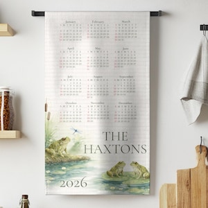 May include: A personalized kitchen towel featuring a 2026 calendar with a frog pond illustration. The towel includes the family name "The Haxtons" and a watercolor image of frogs on lily pads.