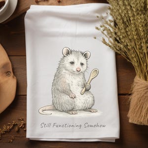 May include: White kitchen towel featuring a watercolor illustration of an opossum holding a wooden spoon. The opossum is gray and white with a pink nose and ears. The text "Still Functioning Somehow" is printed below the opossum.
