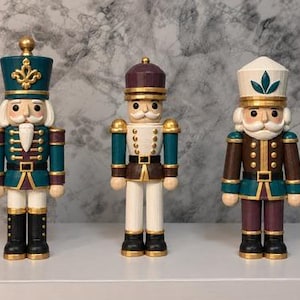 May include: Three decorative nutcracker figurines standing in a row. The left figure wears a teal and gold uniform with a tall hat. The centre figure features a white and teal uniform with a rounded cap. The right figure has a white hat and brown coat.