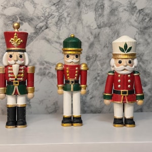 May include: Three nutcracker figurines standing in a row. The left figure wears a red hat with a gold emblem, red jacket, and black boots. The centre figure has a green hat, red jacket, and white trousers. The right figure features a white hat with a green leaf emblem, red jacket, and black boots.