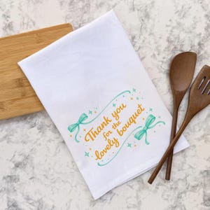 Könnte beinhalten: Weißes Küchentuch mit dem Text "Thank you for the lovely bouquet" in Orange und Türkis. Das Design umfasst türkisfarbene Schleifen und Sterne. Holzlöffel und -spatel liegen neben dem Tuch.