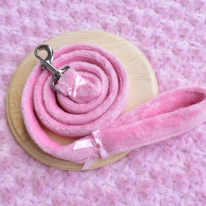 May include: A pink plush dog leash with a silver metal clasp and a matching pink bow. The leash is coiled on a wooden surface, set against a soft, textured pink background. A second pink bow adorns the leash near the handle.