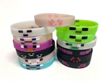 Op de afbeelding: Een stapel kleurrijke siliconen armbanden met pixelontwerpen, geïnspireerd door het populaire videospel Minecraft. De armbanden bevatten verschillende personages en symbolen uit het spel, waaronder een creeper, een varken en een diamant.