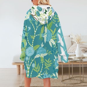 May include: A teal hooded blanket with a floral and bird pattern. The design features large blue flowers, green leaves, and white cranes. The hood has a white floral pattern.