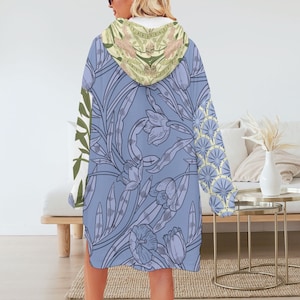 May include: A hooded blanket with a floral design. The blanket features a light blue background with a large floral pattern in shades of purple and gray. The hood has a cream-colored lining and a floral pattern in green and pink.