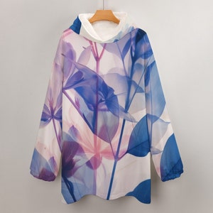 Pode incluir: Uma manta com capuz com um design floral em tons de azul, roxo e rosa. A manta tem um interior branco e fofo e está pendurada num cabide de madeira. O design apresenta folhas e caules translúcidos.