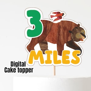 May include: A white cake with a decorative cake topper featuring a brown bear, a green number 3, and the name "MILES" in yellow. The words "Digital Cake topper" are also visible.